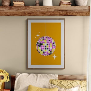May include: A framed print of a disco ball hanging from the ceiling against a yellow background. The disco ball is purple, black, and white. There are two white stars on the background.