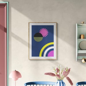 May include: A framed abstract art print with a navy blue background and geometric shapes in yellow, pink, black, and white. The artwork hangs on a cream-colored wall. A pink lamp, blue chairs, and a vase with flowers are in the foreground.