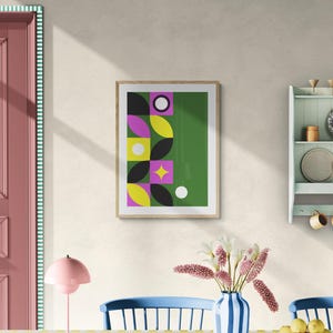 May include: A framed art print with a geometric design featuring black, yellow, pink, and white shapes on a green background. The artwork is displayed on a cream-colored wall in a dining room setting. A pink lamp and a vase with flowers are on the table.