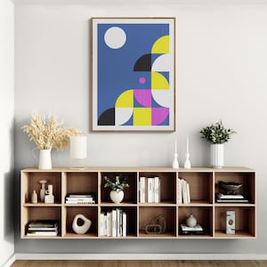 May include: A framed abstract art print with a geometric design in blue, yellow, black, white, and pink hangs above a wooden storage unit. The room is decorated with vases, books, and candles.