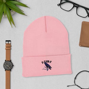 Könnte beinhalten: Hellrosa Beanie mit umgeschlagenem Saum. Auf der Vorderseite des Beanie befindet sich eine dunkelblaue Grafik eines Raben auf einem Ast, darüber der Text "F-CAW-F". Der Beanie ist aus weichem Strickmaterial.
