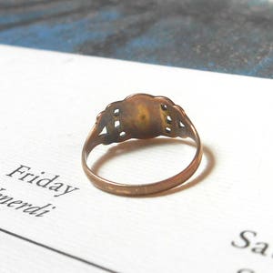 Puede incluir: Un anillo de oro con un dise&ntilde;o decorativo en la parte superior. El anillo est&aacute; sobre una superficie blanca con la palabra "Friday" escrita en ella.