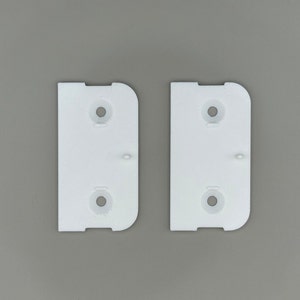Op de afbeelding: Twee witte kunststof beugels met elk drie gaten. De beugels zijn ontworpen om aan een oppervlak te worden bevestigd.