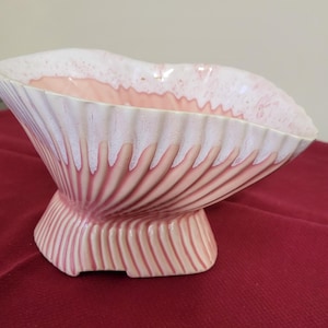 May include: A ceramic decorative bowl with a scalloped design. The bowl is primarily white with pink accents and a textured, ribbed exterior. The bowl sits on a short, pedestal base and is displayed on a red surface.