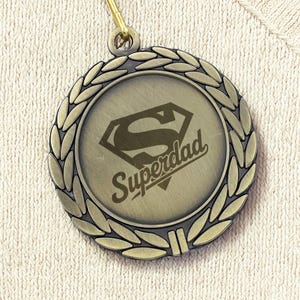 May include: A bronze-colored medal with the word "Superdad" and a Superman logo. The medal is surrounded by a laurel wreath design and hangs from a red, white, and blue striped ribbon.