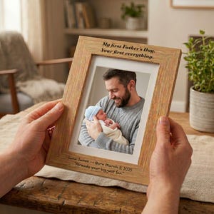 May include: A light oak picture frame holding a photograph of a father and baby. The frame is inscribed with "My first Father's Day. Your first everything." and "Luca James - Born March 3, 2025 Already your biggest fan."
