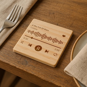 May include: A square wooden coaster with engraved music-themed design. The coaster features the song title "A Sky Full Of Stars" by Coldplay, a waveform, and playback controls. The coaster is on a wooden table next to a fork and a plate.
