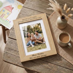 Può includere: Una cornice in legno chiaro con una foto di famiglia. La cornice ha il testo "Any man can be a father, but it takes someone special to be a dad." e "Anne Geddes For our dad, always." Una tazza di caffè e un vaso sono sul tavolo.