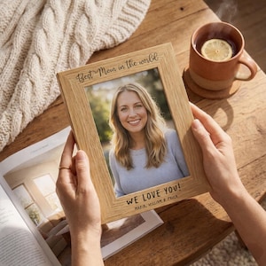 May include: A wooden picture frame with the text "Best Mom in the world" and "WE LOVE YOU!" is held up. The frame contains a photo of a smiling woman. A mug of tea with a lemon slice sits nearby.