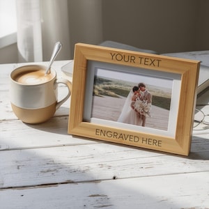 May include: A light brown wooden picture frame with the text "YOUR TEXT" and "ENGRAVED HERE" engraved on the frame. The frame holds a photo of a couple. A mug of coffee sits to the left.