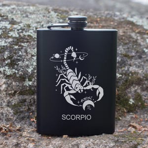 Peut inclure: Une flasque noire en acier inoxydable avec un motif de scorpion blanc, des éléments célestes et le mot "SCORPIO". La flasque a un bouchon à vis et est utilisée pour transporter des boissons alcoolisées. Le design est sur le thème du zodiaque.