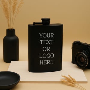 May include: A black metal flask with a silver lid. The flask has the text "YOUR TEXT OR LOGO HERE" engraved on the front.