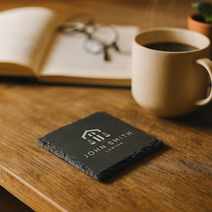 May include: A square, black slate coaster with the text "JOHN SMITH LAWYER" and a graphic of a building and scales. A mug of coffee, an open book, and glasses are in the background, all on a wooden surface.