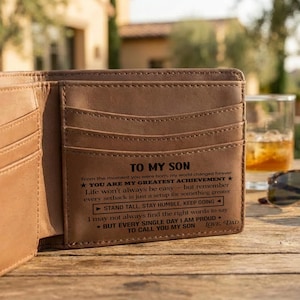 May include: Open brown leather wallet with multiple card slots. The wallet has the text "TO MY SON" and a message from "DAD" printed on the inside. Sunglasses and a glass of amber liquid are in the background.