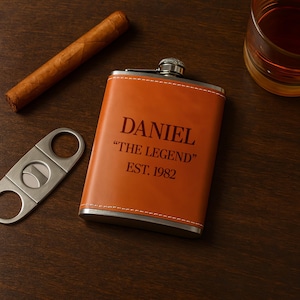 Puede incluir: Un petaca de cuero naranja con el nombre "DANIEL", la frase "THE LEGEND" y el año "EST. 1982" impresos. También hay un cigarro, un cortador de cigarros y un vaso de líquido ámbar.
