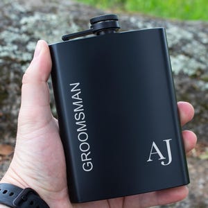 Puede incluir: Una petaca negra con la palabra "GROOMSMAN" impresa verticalmente en blanco. Las iniciales "AJ" están impresas en blanco en la esquina inferior derecha. La petaca tiene una tapa y una bisagra negras. La petaca se sostiene en una mano.