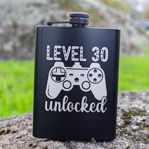 Puede incluir: Petaca negra de acero inoxidable con tapón de rosca. La petaca presenta el texto "LEVEL 30 unlocked" con un gráfico blanco de un mando de videojuegos. La petaca es rectangular y está sobre una roca.