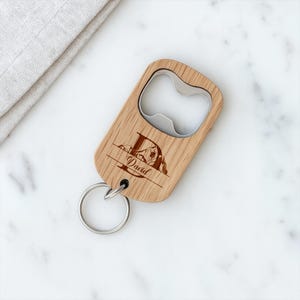 Op de afbeelding: Een houten flesopener met een metalen opener en een sleutelhanger. De opener is gegraveerd met de letter "D", de naam "David" en een berglandschap. Het hout heeft een natuurlijke nerf.