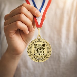 May include: A gold-colored medal with the text "NATHAN WORLD'S BEST BOYFRIEND" is held by a person. The medal hangs from a red, white, and blue ribbon. The medal has a laurel wreath design around the edge.