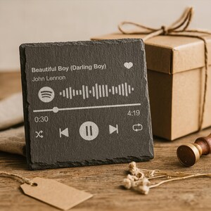 Può includere: Un sottobicchiere quadrato in ardesia grigio scuro con un design dell'interfaccia del lettore musicale. Il sottobicchiere presenta il titolo della canzone "Beautiful Boy (Darling Boy)" di John Lennon, una barra di riproduzione e il logo Spotify. Una confezione regalo e un timbro sono sullo sfondo.