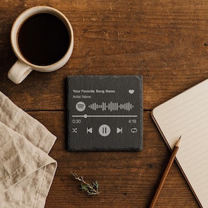 以下が含まれることがあります： 音楽の波形デザインと「Your Favorite Song Name」と「Artist Name」のテキストが入った、正方形の濃い灰色のスレートコースター。コースターには、音楽コントロールアイコンとタイマーも付いています。