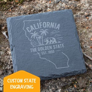 Può includere: Un sottobicchiere quadrato in ardesia grigio scuro con un contorno inciso della California, palme, un orso e il testo "CALIFORNIA THE GOLDEN STATE EST. 1850". Nell'angolo in basso a sinistra c'è uno striscione giallo e arancione con la scritta "CUSTOM STATE ENGRAVING".