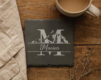 Puede incluir: Un posavasos cuadrado de pizarra gris oscuro con la letra "M" y un diseño de montaña, con el nombre "Macias" debajo. El posavasos está sobre una superficie de madera, junto a un paño beige y una taza de café.