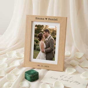 Puede incluir: Un marco de fotos de madera clara con una fotografía de una pareja besándose. El marco está grabado con los nombres "Emma ♥ Daniel" y la fecha "06.14.2026". Pétalos blancos y una pequeña caja verde en primer plano.