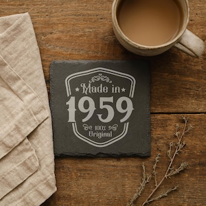 May include: A square, dark gray slate coaster with the text "Made in 1959 100% Original" in white. The coaster is on a wooden surface, next to a beige napkin and a mug of coffee.