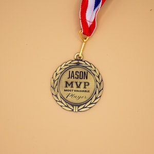 May include: A gold-colored medal with the text "JASON MVP MOST VALUABLE PLAYER" inside a laurel wreath. The medal hangs from a red, white, and blue striped ribbon, suitable for sports awards and recognition.