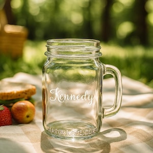 May include: A clear glass mason jar with a handle, the name "Kennedy" in cursive script, and the words "MASON ROME GLASS" at the bottom. The jar sits on a checkered blanket with a strawberry and an apple.