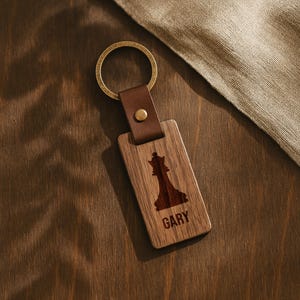 May include: Wooden keyring with a brown leather strap and brass ring. The rectangular wooden piece features a chess queen design and the name "GARY" engraved in a dark brown colour. The keyring is on a dark wooden surface.