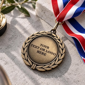 May include: A round bronze-toned medal featuring a laurel wreath border and a central flat area with the text "YOUR TEXT OR LOGO HERE" engraved in black. The medal is attached to a ribbon with red, white, and blue stripes.