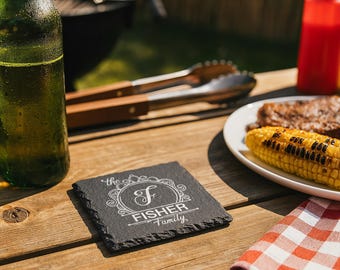 Puede incluir: Un posavasos cuadrado de pizarra negra con el texto "the Fisher Family" en escritura blanca. El posavasos está sobre una mesa de madera, junto a un plato de maíz y costillas a la parrilla, una botella de cerveza y una servilleta a cuadros rojos y blancos. Una barbacoa está en el fondo.