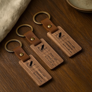 Può includere: Tre portachiavi in legno con cinturini in pelle e anelli in metallo. Ogni portachiavi ha il testo "Drive safe. I need you here with me" e una lettera "A" stilizzata incisa sul legno. I portachiavi sono su una superficie di legno.