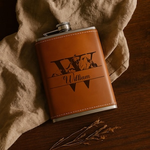 May include: A brown leather flask with a silver cap and trim. The flask features a mountain and letter design with the name "William" engraved on the front. The flask is rectangular and has white stitching.