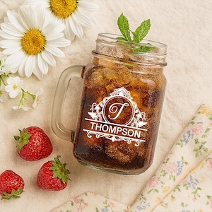 May include: A clear glass mason jar mug filled with a dark beverage and ice, garnished with mint. The mug features a white monogram design with the name "THOMPSON". Strawberries and daisies are arranged around the mug.