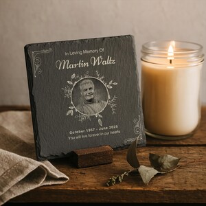 Op de afbeelding: Een zwarte leisteen gedenkplaat met de gegraveerde tekst "In Loving Memory Of Martin Waltz" en een portret. De plaat wordt ondersteund door een klein houten blok. Een brandende kaars in een glazen pot staat rechts.