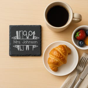 Puede incluir: Un posavasos de pizarra negra con el nombre "Mrs. Johnson" e iconos de artesanía. Una taza de café, un plato con un cruasán y un cuenco pequeño con fruta están sobre una superficie de madera clara.