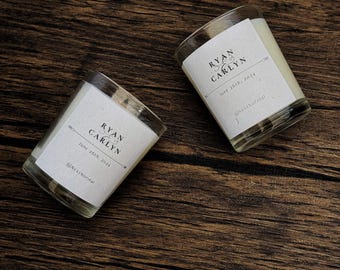 May include: Two small glass candle jars with white labels. The labels have black text that reads "Ryan & Carlynn" and "June 26th, 2024" with the hashtag "@NextNormal".