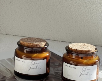 May include: Two amber glass jars with cork lids. Each jar has a white label with the names "Olivia Jonathan" and the date "JUNE | 13 | 2023" printed on it. The jars are likely candles or small containers.