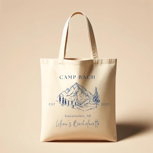 May include: A beige canvas tote bag with a blue mountain scene and the text "Camp Bach Est 2025 Kananaskis, AB  Lilian's Bachelorette" printed on it.