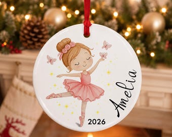 May include: A round white ceramic ornament featuring a cartoon ballerina with brown hair in a bun, wearing a pink tutu and ballet slippers. The design includes small pink butterflies, yellow stars, the name Amelia in black script, and the year 2026.