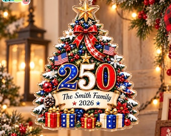 Puede incluir: Un adorno con forma de árbol de Navidad que muestra el número 250, dos banderas estadounidenses y una pancarta con el texto The Smith Family 2026. El diseño incluye lazos rojos, cajas de regalo, piñas y una estrella dorada, colgado de una cinta roja.