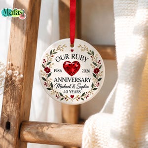 May include: A round white ceramic ornament hanging from a red ribbon. The design features a red faceted heart graphic, green leaves, and red berries. Text reads: OUR RUBY ANNIVERSARY, 1986, 2026, ANNIVERSARY, Michael & Sophia, 40 YEARS.