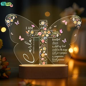 May include: A clear acrylic butterfly-shaped night light with a cross in the center. The cross is decorated with floral designs and the words "I am Carla Baptized 10.12.2024" are written on the left side of the light. The right side of the light has a list of bible verses and phrases, including "beautiful, chosen, a child of God, created in his image, made for a purpose, a conqueror, loved forever".