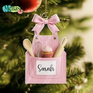 May include: Pink apron-shaped Christmas ornament with a bow, a wooden spoon, whisk, cupcake, and rolling pin. The apron has a pocket with the name "Sarah" on it. The ornament is hanging from a gold string.