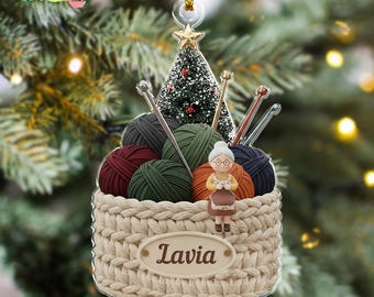 May include: A Christmas ornament featuring a basket filled with yarn balls in shades of burgundy, green, and gray, with knitting needles. A miniature Christmas tree and a small figure of a woman with glasses are also included. The ornament has the name "Lavia" on it.