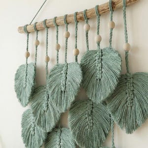 May include: A macrame wall hanging featuring multiple sage green feather-shaped designs suspended from a natural wooden dowel. Each feather is intricately knotted with a textured appearance, and accented with wooden beads. The piece is designed for home decor.