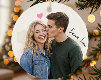 May include: A round, white ceramic ornament with a photo of a smiling couple, one kissing the other's cheek. Two pink hearts are above the couple. The words "Forever Love" are written in black script. A string is attached for hanging.